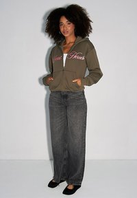 Oliwkowa bluza z zamkiem i różowym napisem na piersi, zestawiona z luźnymi szarymi dżinsami i czarnymi balerinami.