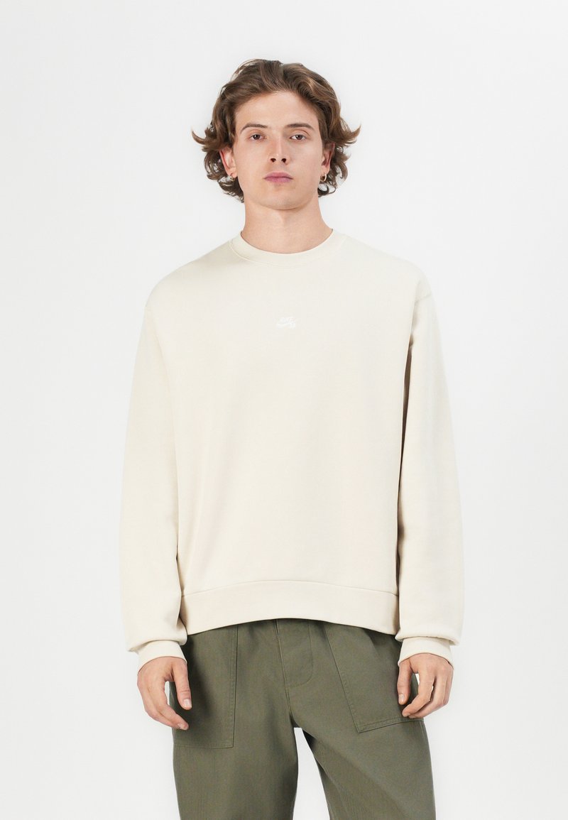 Bež majica s kapuljačom, okruglog izreza i rebrastim rukavima, s malim bijelim logotipom na prsima. Opusteni kroj i glatka tekstura.