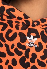 Oranje hoodie met zwarte luipaardvlekken, voorzien van een geborduurd wit Adidas-logo op de borst. Zachte stof met capuchon.