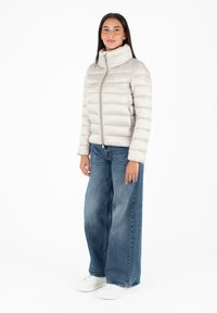 Lichtgrijs pufferjack met hoge kraag, ritssluiting aan de voorkant en horizontaal gestikt patroon. Gepaard met wijde blauwe jeans en witte sneakers.