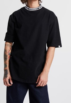 Osoba w czarnej koszulce z podwiniętymi rękawami i białym kołnierzykiem z logo "TOMMY JEANS", pokazująca wytatuowaną lewą rękę.