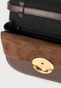 Καφέ σουέντ καπάκι με μεταλλικό χρυσό κλείσιμο, αποκαλύπτοντας ένα σκοτεινό εσωτερικό και μια τσέπη με φερμουάρ, τοποθετημένο σε μια λεία σκούρα καφέ βάση.