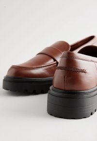 Pruunid nahast mokassiinid, millel on õmmeldud pealiskate, jäme must platvormpõhi ja sile tekstuur. Ees on dekoratiivne rihm.