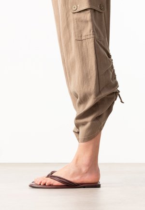 Bare foot wearing brown flat sandals next to cropped beige pants with buttoned pocket and drawstring detail.