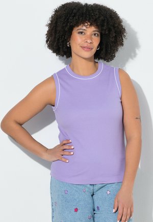 Femme aux cheveux bouclés portant un haut lavande sans manches et un jean bleu clair décoré de patchs étoiles colorés, debout, main sur la hanche.