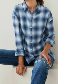 Camicia a scacchi blu con accenti bianchi e navy, dotata di colletto e maniche arrotolate. Indossata con jeans in denim blu strappati.