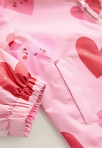 Světle růžová nepromokavá bunda s velkými vzory srdcí v různých odstínech růžové a červené, elastickými manžetami a knoflíkovým detailem.