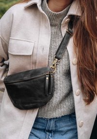 Woman wearing light beige buttoned jacket over gray knit sweater and blue jeans with black leather crossbody bag fastened with a brass clip.