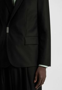 Black tailored blazer with a sleek design, textured fabric, and a single pocket; features a subtle logo on the lapel and contrasting mint-colored cuff.