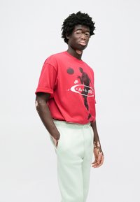 Κόκκινο βαμβακερό t-shirt με γραφική εκτύπωση καλαθοσφαιριστή, συνδυασμένο με ανοιχτό πράσινο παντελόνι φόρμας. Casual, άνετη εφαρμογή.