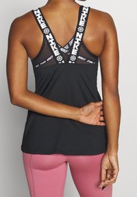Černý sportovní tank top s kontrastními bílými značkovými popruhy, s měkkou síťovinou na zádech a hladkým, lehkým materiálem.