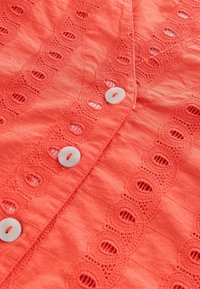 Tissu corail avec couture texturée, orné d'un galon en dentelle et de fermetures à boutons blancs. Le détail comprend des découpes circulaires dans le motif en dentelle.