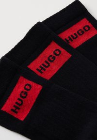 Zwarte sokken met rode rechthoekige panelen waarop het woord "HUGO" in zwarte tekst staat. Zacht materiaal met een geribbelde structuur aan de bovenkant.
