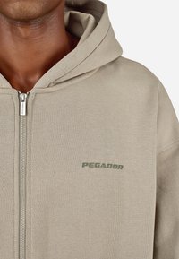 Smėlio spalvos medvilnės mišinio hoodie su užtrauktuku priekyje, priekiniame kišenėje ir žalia „PEGADOR“ logotipo krūtinės srityje.