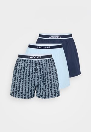 Drei Paare Lacoste Herren-Boxershorts: eines hat ein navyblaues geometrisches Muster, eines ist hellblau, und eines ist ein einfarbiges Navy mit kontrastierenden Taillenbünden.