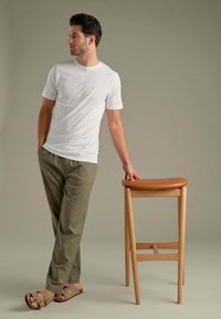 Hvid t-shirt med korte ærmer, olivengrønne bukser og brune sandaler. Læner sig op ad en lys trækstol med et tan polstret sæde.