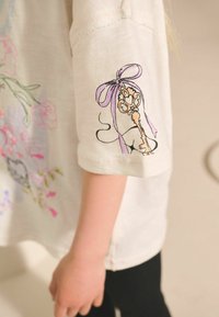T-shirt crème à manches courtes avec un design de clé brodé sur la manche. Présente des imprimés floraux pastel et des détails de couture contrastants.