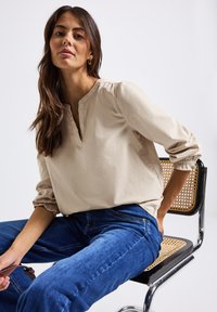 Frau mit langen braunen Haaren, die eine beige Bluse und blaue Jeans trägt, sitzt auf einem Stuhl aus geflochtenem Rohr und Chrom vor einem weißen Hintergrund.