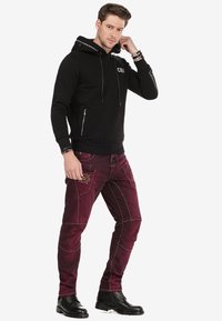 Zwarte hoodie met ritszakken en trekkoorden, gecombineerd met bordeauxrode jeans met geborduurde details en contrasterende stiksels. Zwarte laarzen.