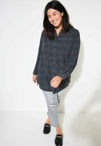 Langärmliges, dunkel gemustertes Hemd mit kleinen bunten Punkten, kombiniert mit hellgrauen, zerrissenen Jeans und schwarzen Slippers mit Kettenakzent.