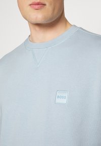 Ljusblå sweatshirt med ribbad rundhals, som har en liten broderad fyrkantig logotyp i matchande färg på vänstra bröstet. Slät textur.