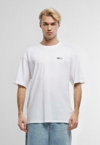 Løs pasform hvid bomulds t-shirt med korte ærmer, rund hals og et lille sort logo på venstre bryst. Glat tekstur.