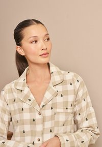 Béžový a bílý kostkovaný pyžamový vršek s malými zelenými vzory stromů, s límečkem, knoflíky a kapsou na levé straně.