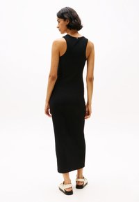 Abito canotta nero a coste con silhouette aderente, orlo fino al ginocchio e dettaglio del tag sul retro. Indossato con sandali leggeri e slip-on.