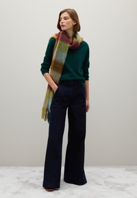 Suéter en verde oscuro, vaqueros de pierna ancha en azul marino y una bufanda de cuadros multicolores con flecos; texturas suaves y contrastes de colores vibrantes.