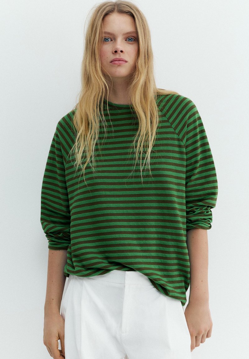 Giovane donna con lunghi capelli biondi che indossa una maglia a maniche lunghe verde e nera a righe e pantaloni bianchi su uno sfondo chiaro e uniforme.