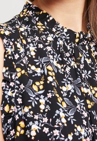 Svart blommig blus med volangkrage, prydd med små rosa, gula och vita blommor på en mörk bakgrund. Mjuk tygtextur.