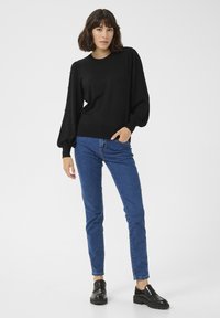 Maglione nero con maniche a sbuffo testurizzate e piccole decorazioni, abbinato a jeans blu skinny e mocassini neri.