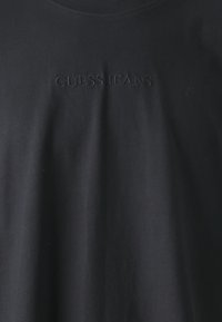 Juodas medvilnės marškinėlis, turintis iškilų „GUESS JEANS“ logotipą priekyje. Audinys atrodo lygus su nedideliu blizgesiu.
