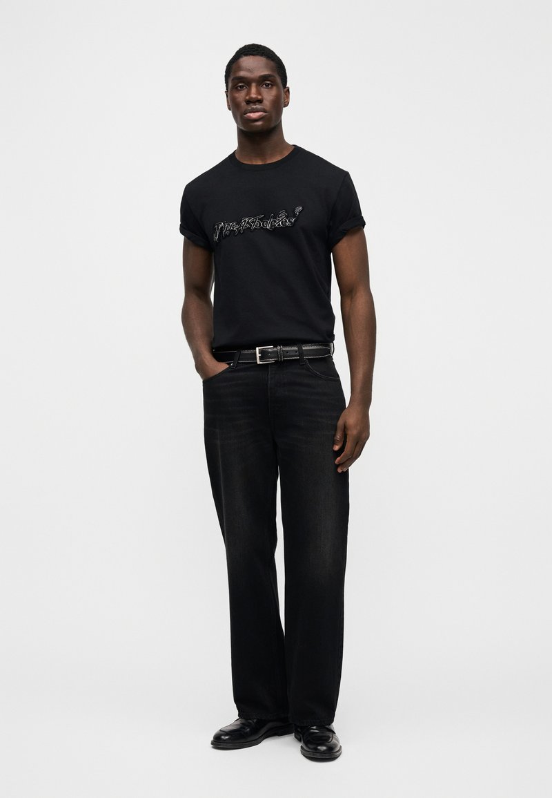 T-shirt en coton noir avec un imprimé graphique sur le devant, assorti à un jean denim noir à jambes larges et une ceinture noire, porté avec des chaussures noires.