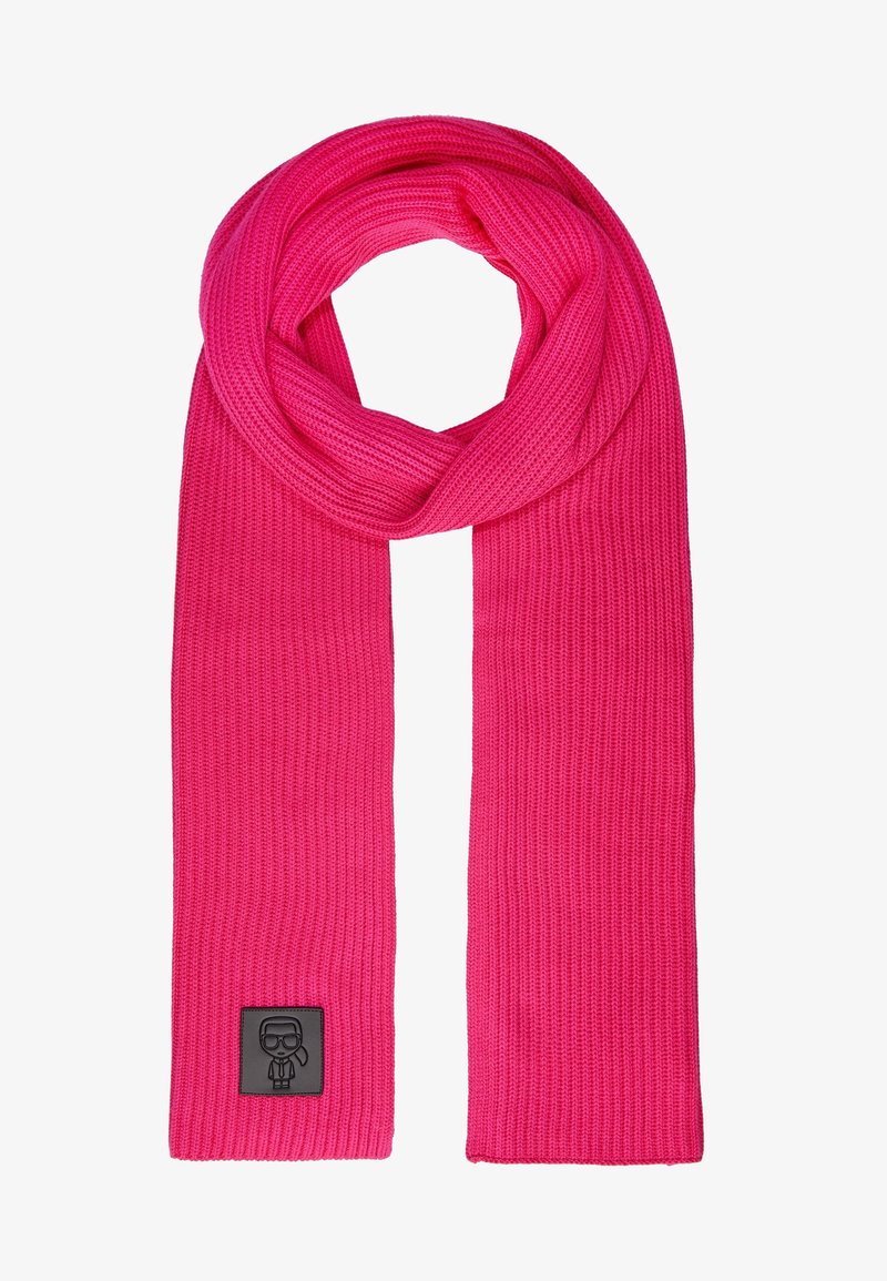 Jasnoróżowy, dziergany szal z prążkowaną fakturą, zawiązany u góry, z małą czarną naszywką przedstawiającą stylizowaną postać w okularach i krawacie.