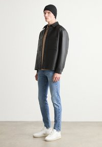 Camisa de cuero negra con dos bolsillos frontales, llevada sobre una camiseta marrón, combinada con vaqueros de un azul claro y zapatillas blancas. Se complementa con un gorro negro.