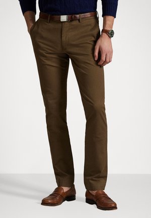 Braune Chinos aus glattem Stoff, mit taillierter Passform, Seitentaschen und einem Gürtel mit einer silbernen Schnalle. Getragen mit braunen Slippern.