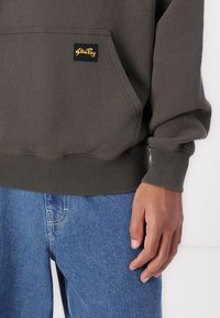 Dark green sweatshirt with a kangaroo pocket, ribbed cuffs, and hem, featuring a yellow logo tag on the pocket. Paired with light blue denim jeans.