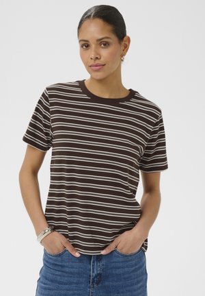 Femme aux cheveux noirs attachés en chignon, porte un T-shirt à rayures horizontales marron et blanc et un jean bleu, debout avec les mains dans les poches.