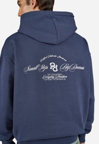 Person wearing a navy hoodie with white text on the back saying "Small Steps Big Dreams" and motivational phrases in cursive font.