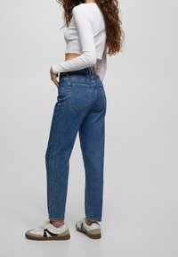 Modré džíny s vysokým pasem a rovnými nohavicemi, vybavené klasickými zadními kapsami a třepeným lemem, kombinované s bílými teniskami a krátkým topem.