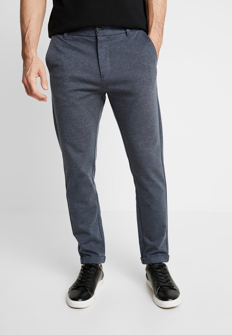 Mies, joka on pukeutunut sinisiin slim fit -vapaa-ajan housuihin, kädet taskuissa ja mustiin lenkkitossuihin, joissa on valkoiset pohjat, valkoista taustaa vasten.