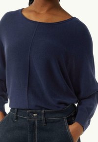 Middengrijze gebreide trui met een brede bootnek, lange dolmanmouwen en een gladde textuur, gecombineerd met donkere denim jeans met contrasterende stiksels.