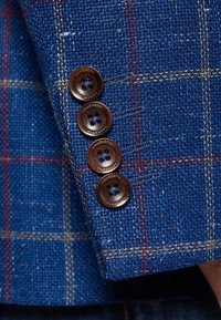 Niebieska tkana tkanina w kratkę, z czterema brązowymi guzikami z etykietą marki. Szczegółowa tekstura i kontrast kolorów.