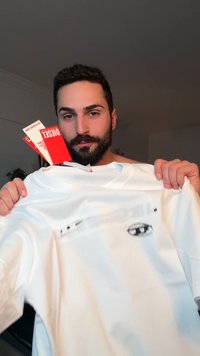 Vīrietis ar bārdu, kas tur baltu T-kreklu ar etiķetēm, priekšpusē neliela melnbalta logo, iekštelpās ar neitrālu fonu.