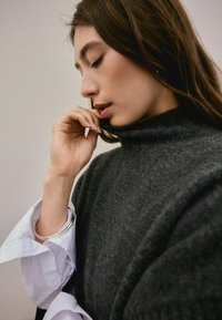 Young woman with long brown hair wearing a dark gray turtleneck sweater and white shirt, touching her lips thoughtfully with her hand.