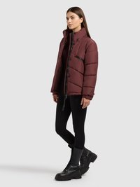 Burgundia värvi paks jakk kõrgema krae, luku ja trukiga sulgemise ning mustade detailidega. Kandmisel mustad retuusid ja massiivsed mustad saapad.