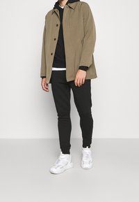 Světlohnědá bunda se zapínáním na knoflíky, pod ní černá mikina s kapucí, černé tepláky a bílé tenisky se vzorovaným detailem.