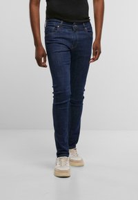 Vaqueros de mezclilla azul oscuro de corte slim, diseñados con cinco bolsillos, sutiles acentos de costura y cierre de cremallera, combinados con zapatillas beige.
