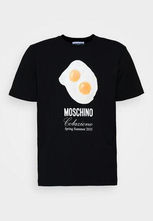 T-shirt nera in cotone con una grafica di uova fritte con due tuorli arancioni. "Moschino Colazione Primavera Estate 2025" stampata in bianco.